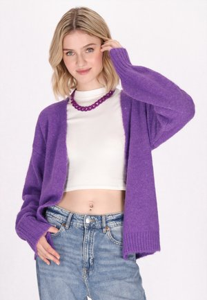 Fialový, nadýchaný kardigan s dlouhými rukávy a žebrovanými manžetami, nošený přes bílý crop top. V kombinaci s modrými džíny s vysokým pasem.