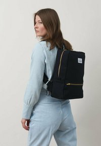 Zaino nero di forma rettangolare, con dettagli in metallo dorato sulla zip e una tasca frontale. Realizzato in tessuto con una finitura opaca. Spallacci attaccati.