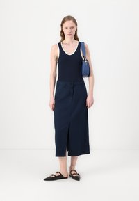 Marineblå ermeløs topp med hvit kant, paret med en lang marineblå skjørt med en frontslitt. Svart slip-on sko med spiss tå. Blå skulderveske.