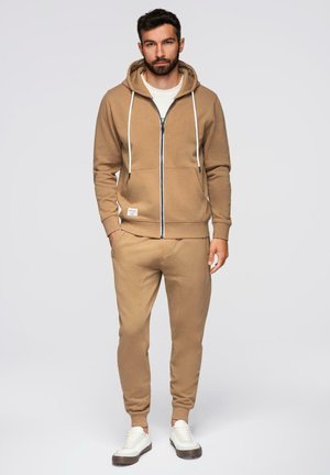 Homme debout portant un sweat à capuche à fermeture éclair marron clair et un pantalon de jogging assorti avec des baskets blanches sur un fond clair.