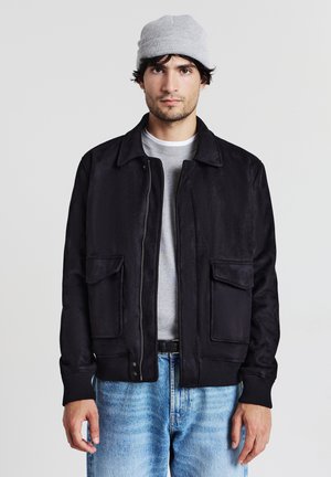 Veste noire zippée avec un col, dotée de deux poches avant, de poignets côtelés et d'une texture lisse, portée par-dessus une chemise grise et un jean clair.