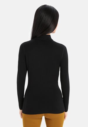 Vrouw met lang donker haar kijkt van ons weg, draagt een aansluitende zwarte coltrui en mosterdgele broek tegen een effen lichte achtergrond.