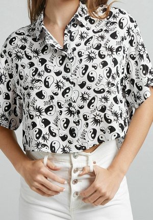 Vrouw draagt een wit kort mouw crop top met zwarte yin-yang symbolen, zonnen en de woorden "love" en "peace," gecombineerd met een witte broek.