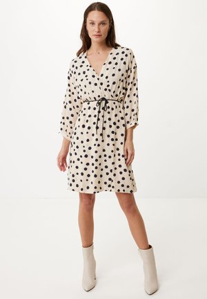 Robe à pois crème avec des pois noirs, décolleté en V, taille ceinturée et manches amples. Portée avec des bottines beige. Texture lisse.