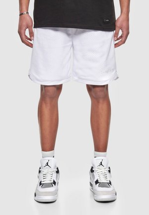 Pantaloni sportivi bianchi con una trama a rete e orlo arrotolato, abbinati a sneakers nere e bianche con un logo prominente e dettagli in rete.
