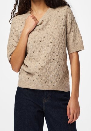 Personne portant un pull tricoté beige à manches courtes avec un motif de feuilles et un jean bleu foncé, tenant le pull près du cou.