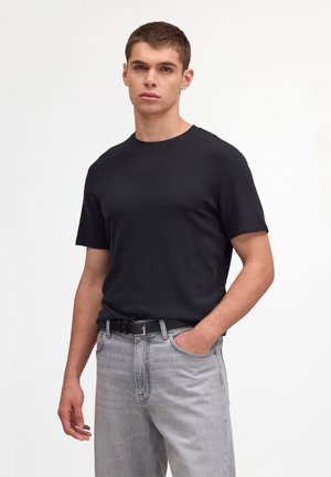 Młody mężczyzna stojący z jedną ręką w kieszeni, ubrany w czarną koszulę z krótkim rękawem, jasnoszare dżinsy i czarny pasek, na neutralnym tle.