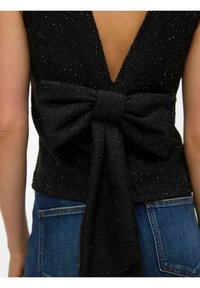 Skats no muguras uz personu, kas valkā spīdošu melnu augšu ar lielu loku, sasietu pie apakšējās muguras, kopā ar zilām džinsa biksēm.