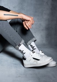 Białe trampki z czarnymi akcentami i wzorem gwiazdy, zestawione z białymi skarpetkami z czarnym napisem, noszone z podwiniętymi szarymi dżinsami.