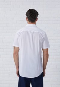 Camisa blanca de manga corta hecha de tela suave, con collar clásico, dobladillo curvado y detalles en el yugo trasero. Diseño simple y limpio.