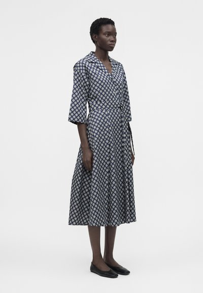 Γυναίκα στέκεται φορώντας ένα μεσομάνικο φόρεμα σε ναυτικό μπλε με λευκά γεωμετρικά σχέδια, μανίκια τριών τετάρτων, κουμπιά στο μπροστινό μέρος και μαύρες μπαλαρίνες.