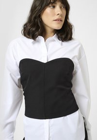 Sort strapless korsettop med en glat overflade, struktureret design og en forlængende underkant over en hvid skjorte.