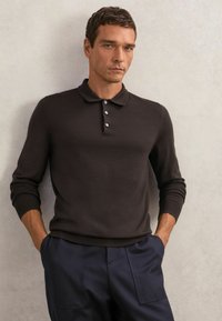 Dunkelbraunes Baumwoll-Poloshirt mit langen Ärmeln, Drei-Knopf-Leiste mit hellen Knöpfen, kombiniert mit marineblauen Hosen.