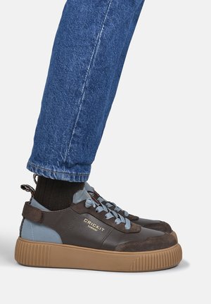 Fod iført brun og blå Crickit Hamburg sneaker med tyk tan sål, sorte sokker og blå cowboybukser mod en ensfarvet lys baggrund.