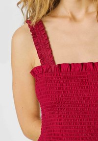 Donna che indossa un abito rosso senza maniche, arricciato, con spalline e scollo arruffati, che mostra spalle e parte superiore del petto.