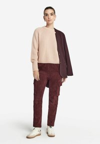 Strickpullover in Hellrosa, kombiniert mit bordeauxfarbenen Wildleder-Cargo-Hosen mit Seitentaschen und weißen Turnschuhen mit braunen Akzenten.