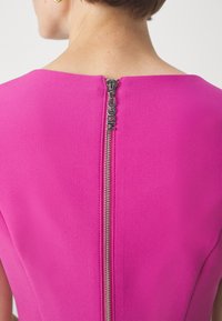 Vue de dos d'une personne portant un haut rose vif avec une fermeture éclair argentée visible courant le long du centre, marquée du mot "HUGO".