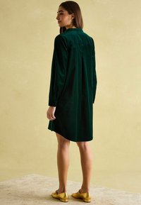 Grünes Samtkleid mit langen Ärmeln, Kragen und gefälteltem Rückendetail, kombiniert mit glänzenden goldenen Schuhen und einem strukturierten beigen Hintergrund.