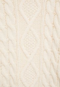 Kermanvärinen neulottu kangas, jossa on palmikkokuvioita ja timanttimaisia muotoja pehmeästä, tekstuurista villamateriaalista.