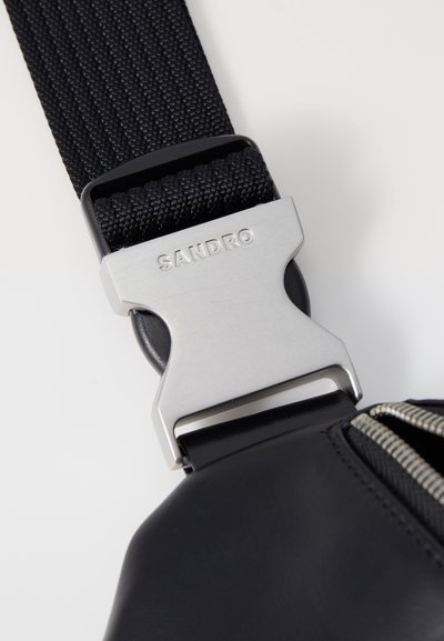 Schwarzes Stoffband mit einer silbernen Metallschnalle, die mit "SANDRO" graviert ist, angebracht an einer schwarzen Tasche mit sichtbarerm Reißverschluss.