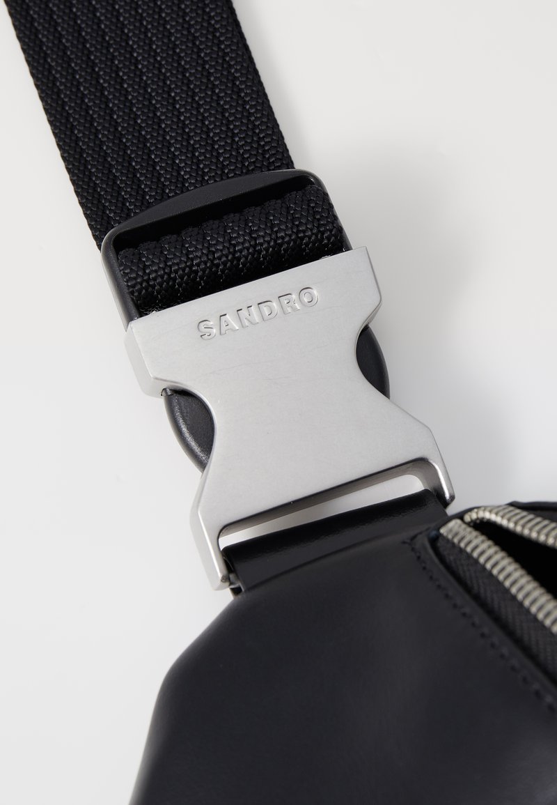 Sangle en tissu noir avec une boucle en métal argenté gravée "SANDRO," attachée à un sac noir avec une fermeture éclair visible.