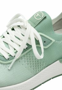 Grüner Sportschuh mit einer strukturierten Mesh-Oberfläche, weißen Schnürsenkeln und einem kleinen Logo-Akzent. Die Sohle ist weiß mit einem leichten grünlichen Farbton.