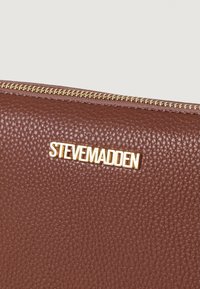 Portofel din piele texturată maro, cu un logo auriu "STEVE MADDEN" și un fermoar auriu pe marginea superioară.