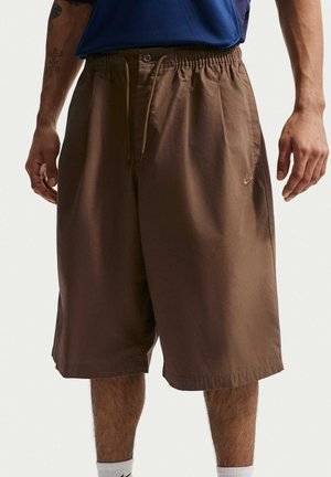 Braune locker sitzende knielange Shorts mit elastischem Bund und Kordelzug, getragen von einer Person mit blauem Hemd und weißen Socken, stehend.