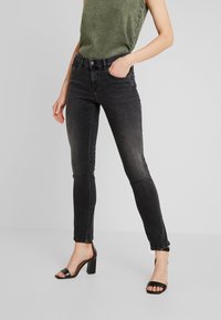 Mujer vestida con jeans skinny negros desgastados, top sin mangas verde oliva y sandalias de tacón negro, de pie con una mano en el bolsillo.