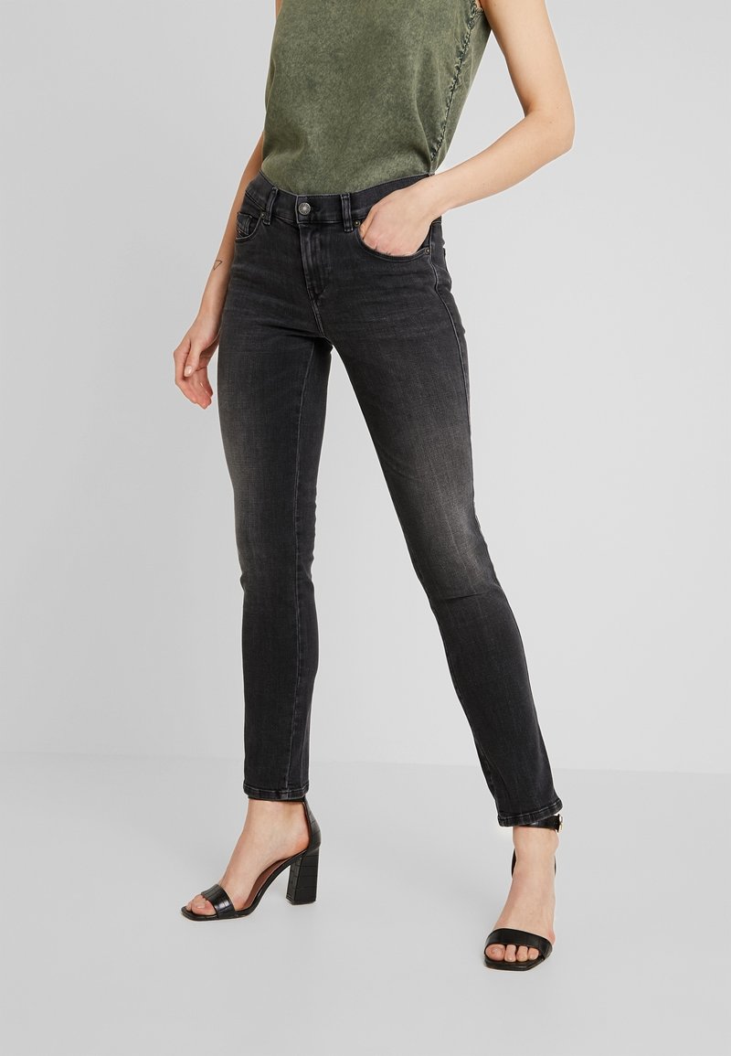 Mujer vestida con jeans skinny negros desgastados, top sin mangas verde oliva y sandalias de tacón negro, de pie con una mano en el bolsillo.