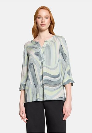 Vrouw draagt een losse blouse met lange mouwen met abstracte groene, grijze en witte golvende patronen, gecombineerd met een zwarte broek.
