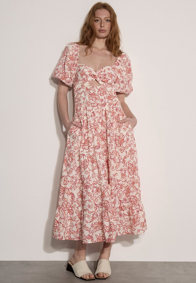 Femme portant une robe midi blanche à imprimé floral rouge, manches bouffantes et poches latérales, associée à des sandales beiges à talon bloc et à bout ouvert.