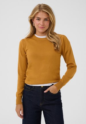 Jeune femme aux longs cheveux blonds portant un pull moutarde et un jean foncé, debout avec une main dans la poche devant un fond gris.