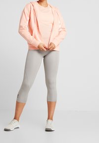 Felpa zip-up rosa chiaro con strisce bianche, abbinata a leggings grigi chiari e sneakers neutre. Abbigliamento sportivo casual.
