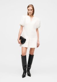 Vestido branco com mangas bufantes, tecido texturizado, decote em V profundo, combinado com clutch preta e botas de couro pretas até ao joelho. Acabamento liso e silhueta definida.