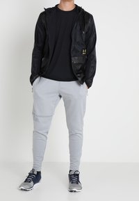 Svart lättvikts zip-up jacka med huva, tillsammans med grå slim-fit joggingbyxor och mörka sneakers. Har framträdande logotyper och dragkedjor.