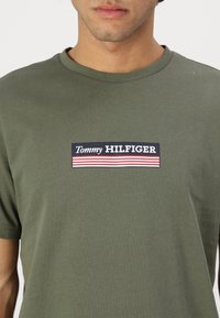 Olive green cotton T-shirt with round neckline; features a rectangular brand logo in navy, white, and red across the chest.