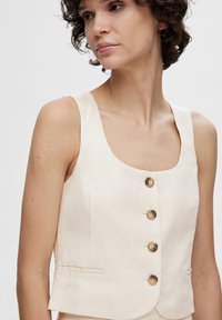 Lys beige ærmeløs vest med en rund halsudskæring, der har fem bronzefarvede knapper og en lille sidelomme. Glat stoftekstur.