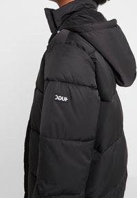 Giacca a vento nera con cappuccio, caratterizzata da un design trapuntato, una texture liscia e un piccolo logo sulla manica sinistra.