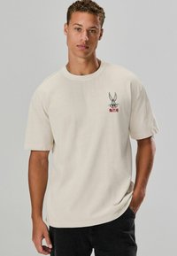 Beige Baumwoll-T-Shirt mit kurzen Ärmeln und Rundhalsausschnitt, mit einem schwarzen und roten Bugs Bunny-Graphic auf der linken Brustseite.
