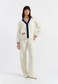 Cremfarbener Cardigan mit marineblauen Akzenten, großen Knöpfen und einer Fronttasche mit gesticktem Design. Kombiniert mit creamfarbenen, weit geschnittenen Hosen.
