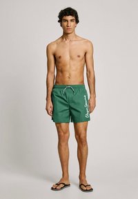 Πράσινα μαγιό σορτς με ρυθμιζόμενο κορδόνι στη μέση, με λευκό λογότυπο "Pepe Jeans" στο πλάι. Το μοντέλο είναι ξυπόλητο, φοράει μαύρα σαγιονάρες.