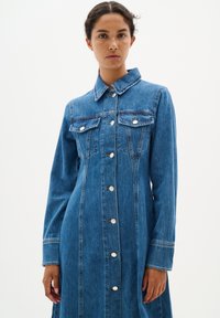 Robe chemise en denim bleu moyen, avec un col, des manches longues, deux poches poitrine et des fermetures à boutons sur le devant avec des accents argentés.