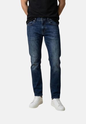 Homme portant un jean slim bleu foncé et des baskets blanches debout, les mains derrière le dos, sur un fond uni.