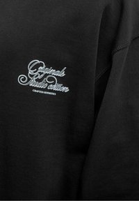 Tissu noir avec texte en cursive blanche « Originals Studio edition » et imprimé plus petit « Crafted Aesthetics » brodés sur la zone de la poitrine.