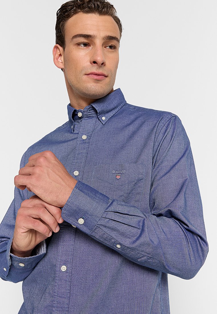 Camisa de botonadura en tejido chambray azul medio, con cuello tradicional, mangas largas y un bolsillo en el pecho con bordado de logo.