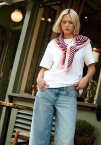 T-shirt blanc associé à un jean large bleu clair, avec un pull en tricot à carreaux roses et noirs drapé sur les épaules.