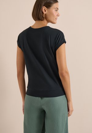 Donna con capelli corti castani vista di profilo, indossa una maglietta nera a maniche corte e pantaloni verdi su uno sfondo beige uniforme.