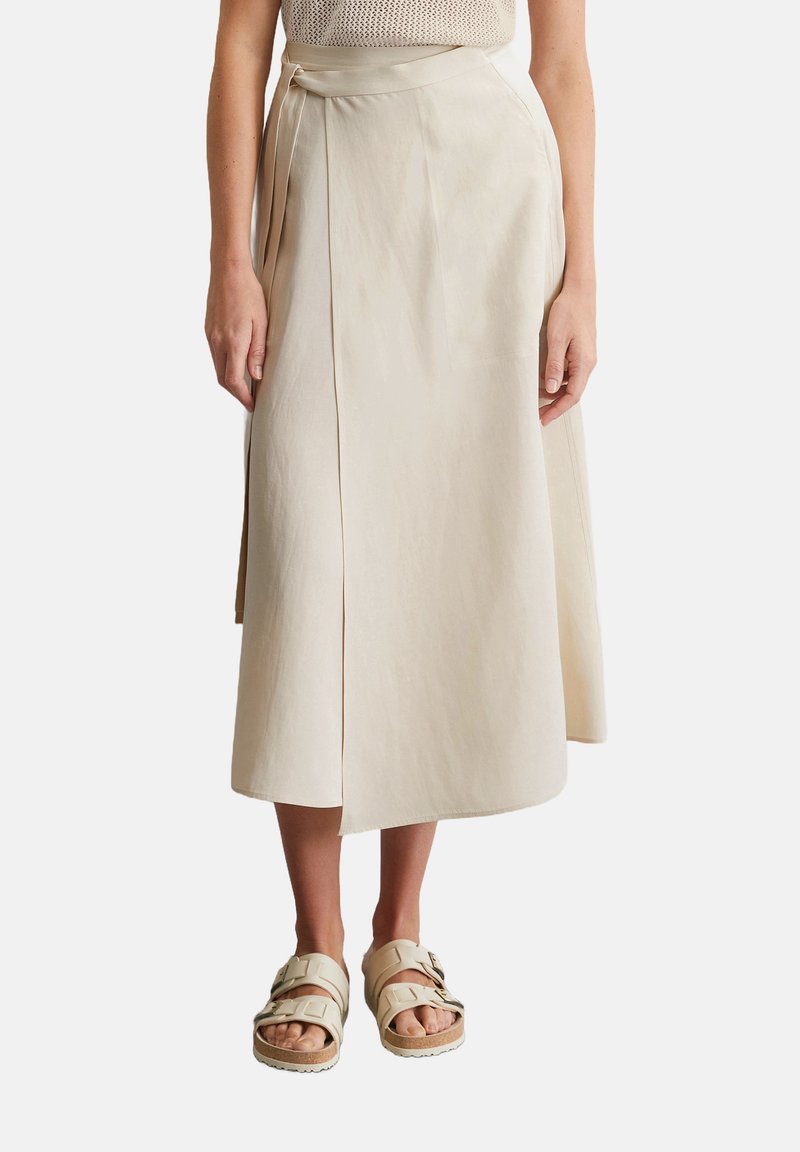 Beige, knee-length A-line skirt in a lightweight fabric, featuring a wrap design and side tie, paired with light-colored strappy sandals.