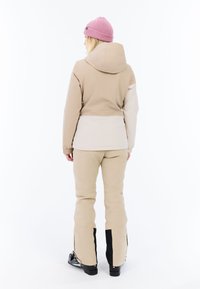 Beige Skijacke mit Kapuze und elastischen Bündchen, kombiniert mit passenden beigen Hosen, die an den Knöcheln mit Reißverschlüssen und schwarzen Akzenten an den Stiefeln ausgestattet sind.